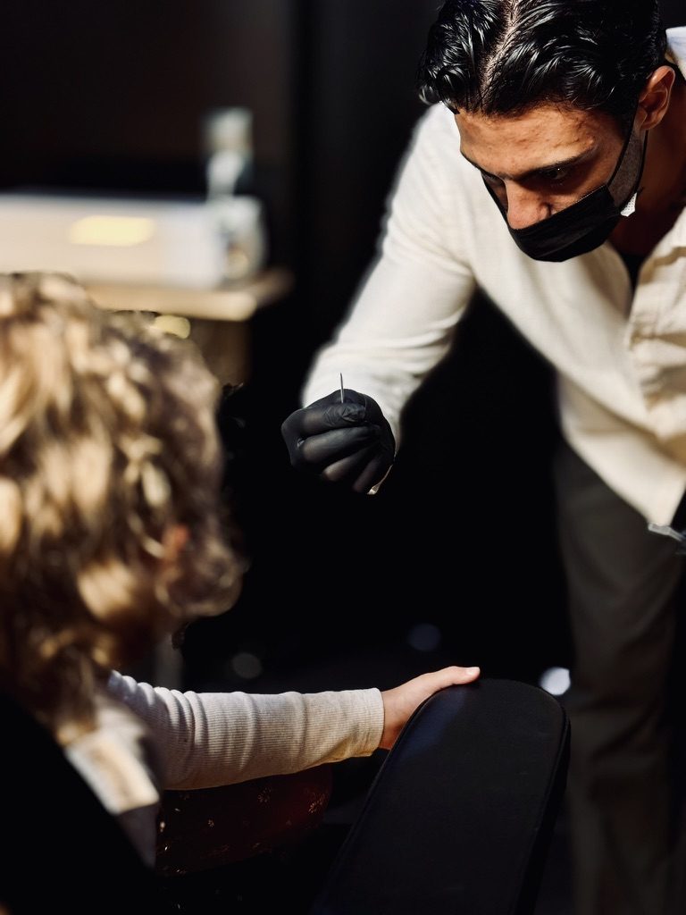Piercer mit Nadel, ein Kind hält sich am Stuhl fest